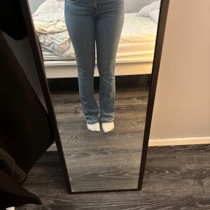 Blå bootcut jeans med rå kant - Säljer ett par ljusblå bootcut jeans med hög midja och rå, fransig kant nertill. Jeansen har klassisk femficksdesign och sitter snyggt över höften och låren innan de släpper ut vid vaden. Perfekta till sneakers eller boots.