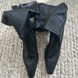 Svarta höga stövlar med klack - Snygga svarta stövlar från Zara i skinn med spetsig tå och låg klack. Modellen är knähög och har dragkedja på insidan för enkel påtagning. Perfekta för att lyfta din outfit med en stilren vibe. Storlek 41 men passar som 40.