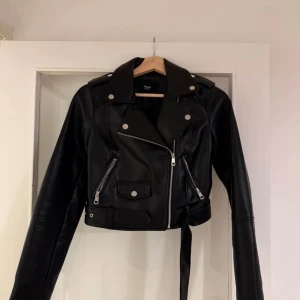 Svart skinnjacka med bälte  - Jackan har silvriga dragkedjor, knappar och ett bälte i midjan. Croppad modell med långa ärmar och coola detaljer som ger en edgy vibe.