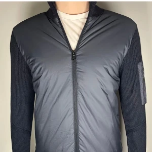 Mörkblå cardigan från J.Lindeberg - Snygg mörkgrå hybridjacka från J.Lindeberg med stickade ärmar och vadderad front. Jackan har hel dragkedja, ribbade detaljer och en praktisk ficka med dragkedja på ärmen. Perfekt för dig som gillar stilren och sportig look.