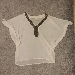 Vit blus från Vero Moda - Säljer en elegant vit blus från Vero Moda med v-ringning och dekorativa pärlor längs halsringningen. Perfekt för en stilren look. Genomskinlig, så perfekt att ha över ett linne. ✨️ finns även i svart