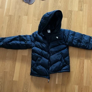 Svart dunjacka från Nike - Säljer en svart dunjacka från Nike med huva och dragkedja. Jackan har en pufferdesign som ger extra värme och stil. Perfekt för kyliga dagar. Storlek 147-158cm. Pris kan diskuteras
