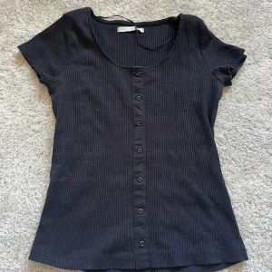 Svart ribbad topp med knappar - Säljer en stilren svart ribbad topp med knappar framtill. Toppen har korta ärmar och en rund halsringning. Perfekt för en avslappnad look💕