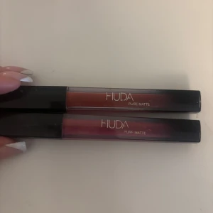 Matta läppstift från Huda - Två matta läppstift från Huda i nyanser av rödbrun och mörkrosa. De har en elegant, långsmal förpackning med svart lock och genomskinlig kropp som visar färgen. Perfekt för en sofistikerad look.