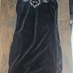Svart sammet klänning från Juicy Couture - Snygg svart sammet klänning från Juicy Couture med glittrande detaljer på bröstet. Klänningen har en unik snörning på sidan som ger en extra touch. Perfekt för en kväll ute eller en speciell tillställning.