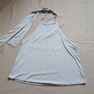 Grå ärmlös halterneck-topp - Säljer en stilren grå halterneck-topp med knytband i nacken. Toppen är ärmlös och tillverkad i en mjuk blandning av viskos och polyester. Supersnygg på fest som till vardags! Eventuell missfärgning som en liten ljusare fläck därav priset. Köpt för ca 250 från Monki. Bortklippt lapp med storlek M o passar S-M beroende på passform 