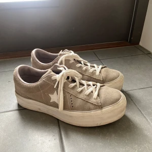 Beige sneakers från Converse - Säljer ett par snygga beige sneakers från Converse i mocka med vit stjärndetalj på sidan. Skorna har snörning och en bekväm, rund tå. Perfekta för en avslappnad stil.