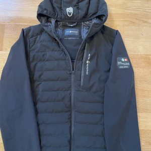 Svart pufferjacka från Sail Racing - Snygg svart pufferjacka från Sail Racing med huva och dragkedja. Jackan har en cool patch på ärmen med texten 'Sail Antarctica Expedition'. Perfekt för kyliga dagar.