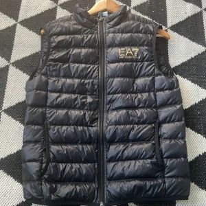 Svart dunväst från EA7 - Snygg svart dunväst från EA7 med dragkedja framtill. Perfekt för kyligare dagar och ger en sportig look. Västen har en quiltad design och är lätt att matcha med olika outfits. 