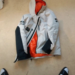 Sailracing thermolite - Säljer en cool vit och svart jacka med orange foder från Gore-Tex. Jackan har en dragkedja och är långärmad med flera logotyper på ärmarna. Perfekt för kyliga dagar med sitt Thermolite-foder.