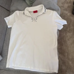 Hugo Pike - Säljer en stilren vit kortärmad pike från Hugo Boss. T-shirten har en klassisk krage med subtila svarta detaljer och en dragkedja framtill. Perfekt för en avslappnad men ändå elegant look.