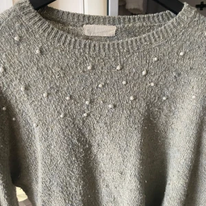 Grå stickad tröja med pärlor - Säljer en stilren grå stickad tröja dekorerad med pärlor på framsidan. Tröjan har en rund halsringning och långa ärmar, perfekt för kyligare dagar. Ett elegant val för den som vill ha något extra i garderoben.