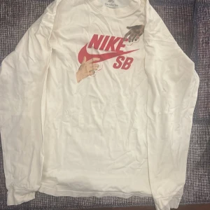 Vit långärmad tröja från Nike SB - Säljer en vit långärmad tröja från Nike SB med ett coolt tryck i rött på framsidan. Perfekt för en avslappnad stil. Tröjan är gjord i bomull och har en bekväm passform.