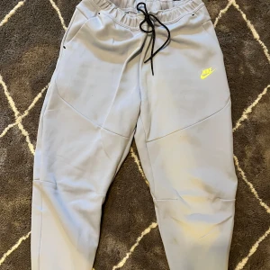 Nike tech byxor - Säljer ett par grå mjukisbyxor från Nike med elastisk midja och snörning. Byxorna har en gul Nike-logga på framsidan och dragkedja på fickan. Perfekta för en avslappnad stil.