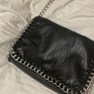 Svart axelväska från Tiamo - Snygg svart axelväska i skinn från Tiamo med silverfärgade kedjedetaljer. Väskan har en stilren design med en dragkedja för säker förvaring. Perfekt för att ge din outfit en edgy touch. Färgen på kedjan har på några ställen bleknat. Hör av dig om du funderar på något!💗