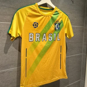 Brasil fotbollströja - Säljer en gul Brasil fotbollströja med gröna detaljer och texten 'BRASIL' på framsidan. Tröjan har korta ärmar och ett tryckt emblem på bröstet. Perfekt för fotbollsfans!