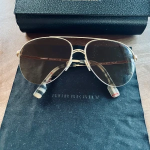 Solglasögon från Burberry - Snygga solglasögon från Burberry med guldfärgad metallram och mörka glas. De har en klassisk pilotform och stilrena detaljer. Perfekta för en elegant look. Nypris ca 2 000kr