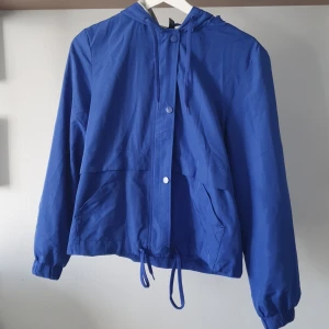 Blå vindjacka från H&M Divided - Säljer en blå vindjacka från H&M Divided med huva och dragsko i midjan. Jackan har dragkedja och knappar framtill samt två stora fickor. Mjukt och vindtätt material! 💙