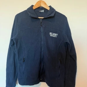 Mörkblå fleecejacka från Columbia - Säljer en mörkblå fleecejacka från Columbia med dragkedja och broderad text 'Belmont University' på bröstet. Jackan har långa ärmar och två fickor med dragkedjor. Perfekt för kyliga dagar!