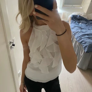 Vit ärmlös blus med volanger - Säljer en elegant vit ärmlös blus med volanger framtill. Blusen har en knytning i nacken och är perfekt för en stilren look. Materialet är lätt och luftigt, vilket gör den bekväm att bära❤️ Perfekt till sommaren💕