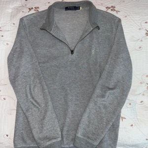 Grå tröja med dragkedja från Polo Ralph Lauren - Säljer en stilren grå tröja från Polo Ralph Lauren med dragkedja vid halsen. Tröjan har långa ärmar och en liten broderad logga på bröstet. Perfekt för en avslappnad och snygg look.