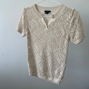 Beige stickad kortärmad tröja från Lindex  - Säljer en beige stickad tröja från Lindex  med korta ärmar och ett snyggt mönster. Tröjan har en rund halsringning med knappar framtill. Perfekt för en stilren och avslappnad look.