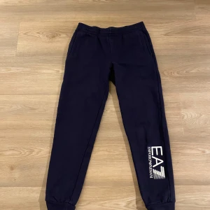 Mörkblå mjukisbyxor från Emporio Armani - Snygga mörkblå mjukisbyxor från Emporio Armani med elastisk midja och muddar vid bensluten. Byxorna har en vit logotyp på benet som ger en stilren look.