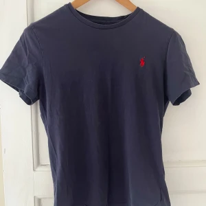 Mörkblå t-shirt från Polo Ralph Lauren - Säljer en stilren mörkblå t-shirt från Polo Ralph Lauren med röd broderad logga på bröstet. T-shirten har en slim fit passform och är tillverkad i mjuk bomull. Perfekt för en avslappnad och snygg look.