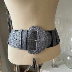 Grått bälte med krokodilmönster 75 cm  - Snyggt grått bälte med krokodilmönster och stor rund spänne. Perfekt för att ge en edgy touch till din outfit. Bältet har en bred design med elastisk baksida för extra komfort. Det svarta är gummi 