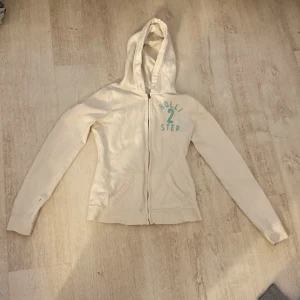 Vit hoodie från Hollister - Säljer en vit hoodie från Hollister med dragkedja och turkos text på bröstet. Den har en mysig huva och långa ärmar, perfekt för en avslappnad stil. Hör av dig vid intresse!