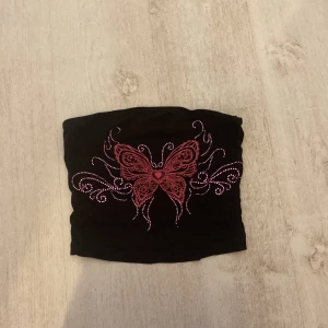 Svart tubtopp med fjärilsdesign - Snygg svart tubtopp med en stor rosa fjäril och dekorativa mönster i rosa och vitt. Perfekt för en cool och unik stil. Passar bra till både jeans och kjol.