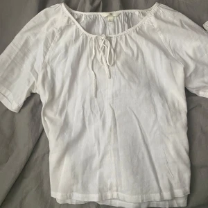 Vit blus med knytband - Säljer en söt vit blus aktig topp med korta ärmar och knytband vid halsen. Perfekt för en avslappnad sommarlook. Blusen har en luftig passform och är gjord i ett lätt material.