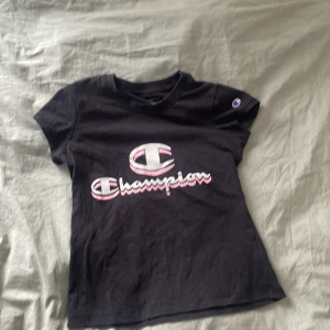 Svart t-shirt från Champion - Snygg svart t-shirt från Champion med deras ikoniska logga i vitt och rosa på bröstet. Perfekt för en casual look. Kortärmad och i mjukt material för skön komfort.