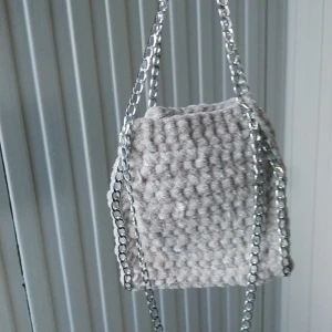 Ljust beigegrå virkad väska med kedja - Snygg  ljus beigegrå väska i virkat material med en cool kedja. Perfekt för att ge din outfit en unik touch. Väskan har en mjuk och mysig känsla.