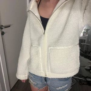 Vit teddyjacka med dragkedja - Mysig vit teddyjacka med dragkedja framtill och långa ärmar. Perfekt för kyligare dagar när du vill hålla dig varm och bekväm. Jackan har en avslappnad passform och två praktiska fickor framtill. Ifrån lager157 o slutsåld 