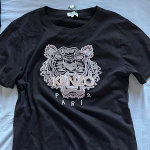 Svart t-shirt med tigerbroderi från Kenzo - Snygg svart t-shirt från Kenzo med ett stort, detaljerat tigerbroderi i silver och vitt på framsidan. Perfekt för dig som vill ha en stilren men ändå iögonfallande look. T-shirten har en klassisk passform och rund halsringning.