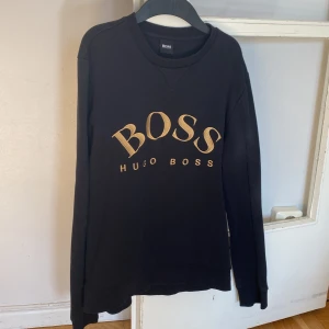 Svart tröja från Hugo Boss - Säljer en stilren svart tröja från Hugo Boss med deras ikoniska logga i guld på bröstet. Tröjan har långa ärmar och en klassisk rund halsringning. Perfekt för en avslappnad men ändå elegant look👍🏻