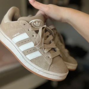 Beige Adidas Campus sneakers - Säljer ett par snygga beige Adidas Campus sneakers med vita detaljer. Skorna kommer men snören i samma färg som skon men skickar även med ett par vita lite tunnare snören. Skorna har inte använt så mycket💗