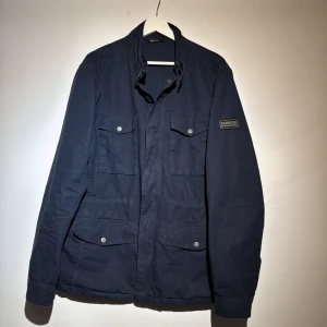 Mörkblå jacka från Barbour - Säljer en stilren mörkblå jacka från Barbour med flera praktiska fickor och knappar. Jackan har en klassisk design med lång ärm och en diskret logga på ärmen. Perfekt för en snygg och funktionell look.