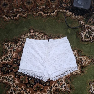 Vita spetsdetaljerade shorts - Säljer ett par vita shorts med blommigt mönster och spetsdetaljer vid bensluten. Shortsen har en dragkedja bak och är perfekta för sommarens varma dagar.