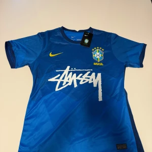 Blå Brasilien fotbollströja från Nike - Snygg blå fotbollströja från Nike med Brasilien-emblem och Stussy-logga. Tröjan har korta ärmar och är tillverkad i Dri-FIT-material för optimal komfort. Perfekt för fotbollsfans som vill ha stil och funktion.