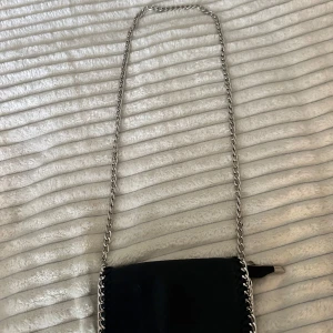 Svart axelväska från Tiamo - Snygg svart axelväska från Tiamo med kedjeaxelrem och dragkedja. Endast lite rost på knapparna annars nästan som ny🥰