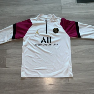 Vit fotbollströja med vinröda detaljer från Jordan - Säljer en vit fotbollströja med vinröda och svarta detaljer från Jordan. Tröjan har en halv dragkedja framtill och ett tryck med texten 'Accor Live Limitless'. Perfekt för fotbollsfans som vill ha en stilren look.