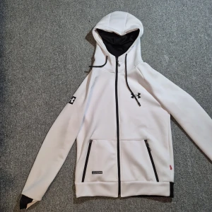 Vit zip hoodie från Under Armour  - Snygg vit vindjacka från Under Armour med svarta detaljer, huva och dragkedja framtill. Jackan har två fickor med dragkedja och logga på bröstet och ärmslutet. Perfekt för dig som gillar sportig och clean stil.