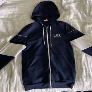  EA7 zip hoodie - Snygg marinblå hoodie från EA7 Emporio Armani med vita detaljer på ärmarna och vit dragkedja. Tröjan har huva med snörning och två fickor framtill. Perfekt för en sportig och stilren look.