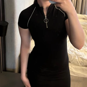 Svart kortärmad klänning med zip - Svart, tight kortklänning från H&M med vita detaljer längs axlarna och en cool dragkedja med ring framtill.  Använd typ 1 gång 
