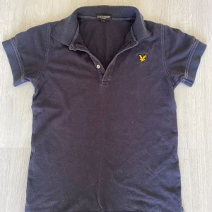 Mörkblå pikétröja från Lyle & Scott - Snygg mörkblå pikétröja från Lyle & Scott med klassisk krage och korta ärmar. Tröjan har gul broderad logga på bröstet och två knappar vid halsen. Tillverkad i mjuk bomull, perfekt för en chill och stilren look.