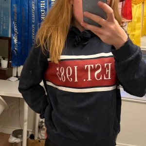 Mörkblå hoodie med EST. 1987-tryck - Säljer en mörkblå hoodie med vit och röd panel framtill och stort EST. 1987-tryck. Tröjan har huva med snörning och långa ärmar. Perfekt för en chill och avslappnad stil.
