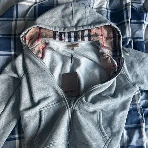 Grå hoodie från Burberry med rutigt foder - Säljer en grå hoodie från Burberry med klassiskt rutigt foder i huvan och broderad logga på bröstet. Jackan har dragkedja och känguruficka framtill. Tillverkad i mjuk bomull med elastan för extra komfort.