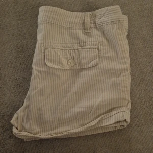 Randiga shorts - Snygga beige/vita shorts från Cubus med tunna ränder. Bakfickor med knapp och uppvikta benslut. 98% bomull, 2% elastan. Låg midja.
